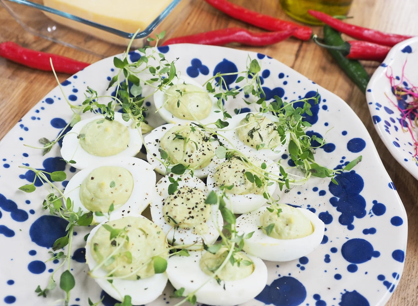 Połówki jajek z zielonym kremem, udekorowane świeżymi ziołami i oprószone pieprzem, ułożone na biało-niebieskim talerzu, w tle czerwone i zielone papryczki chili.