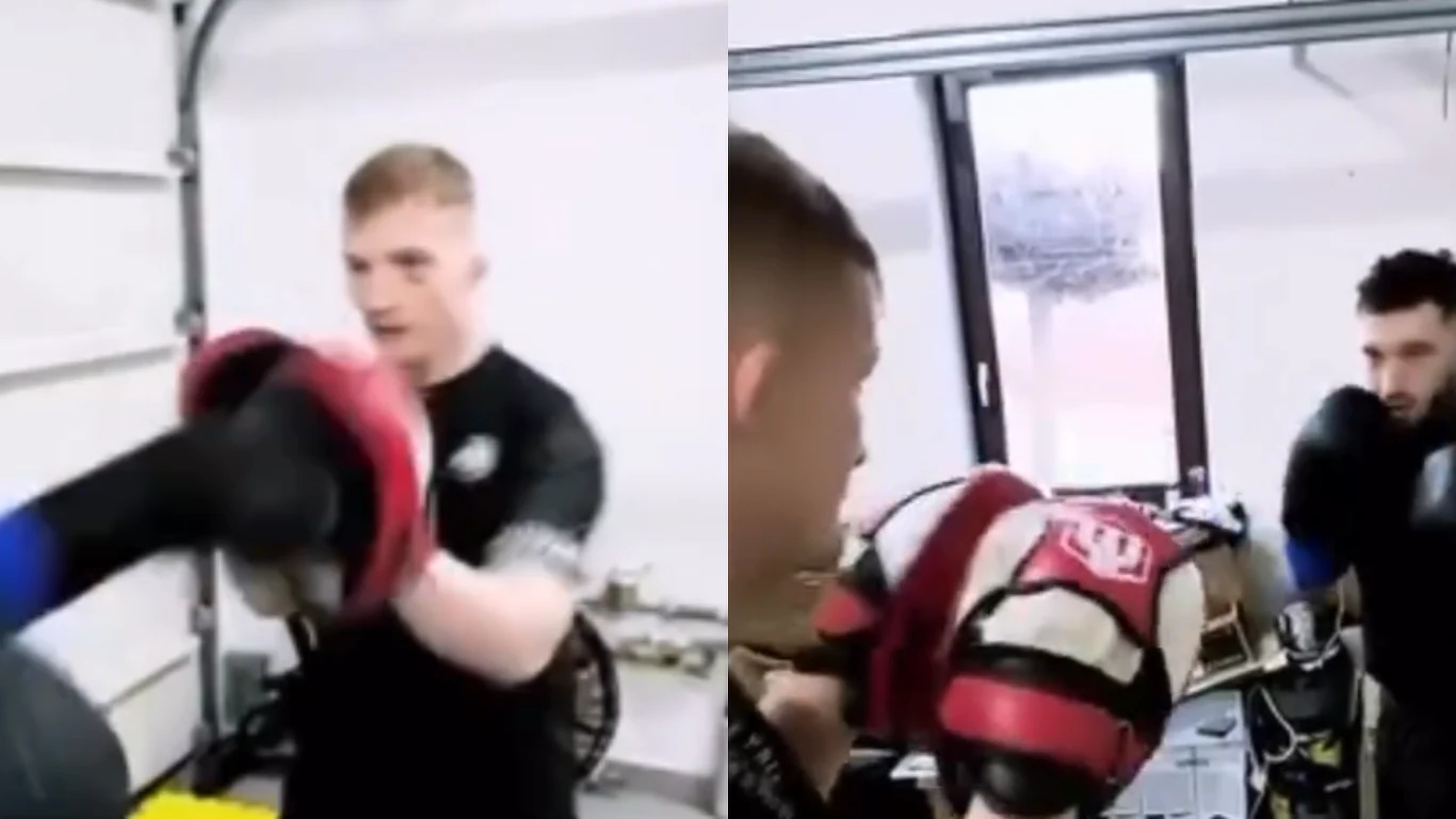Dwóch mężczyzn trenuje boks w zamkniętym pomieszczeniu, jeden z nich uderza w tarcze trzymane przez drugiego, obaj skoncentrowani na ćwiczeniach.