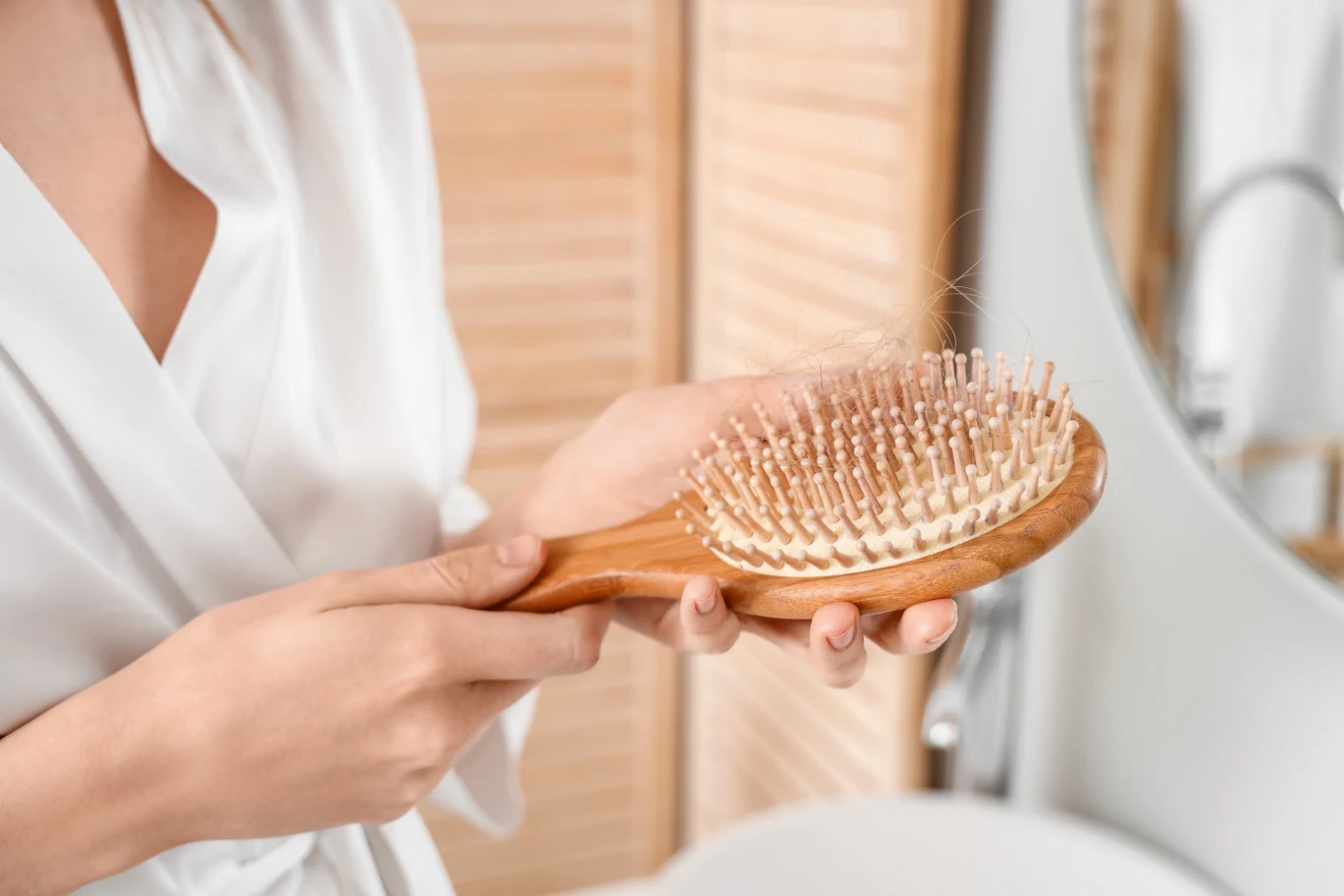 Dłoń trzymająca drewnianą szczotkę do włosów z widocznymi włosami zaplątanymi w jej ząbki, w tle jasna łazienka z lustrem.