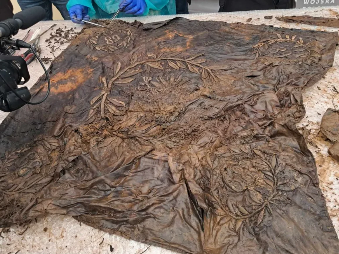 silnie zniszczony, zabłocony sztandar wojskowy z widocznymi śladami zawiłych haftowanych motywów roślinnych, liczbą 89 w rogach oraz otaczającymi przedmiot osobami w rękawiczkach prowadzącymi prace konserwatorskie