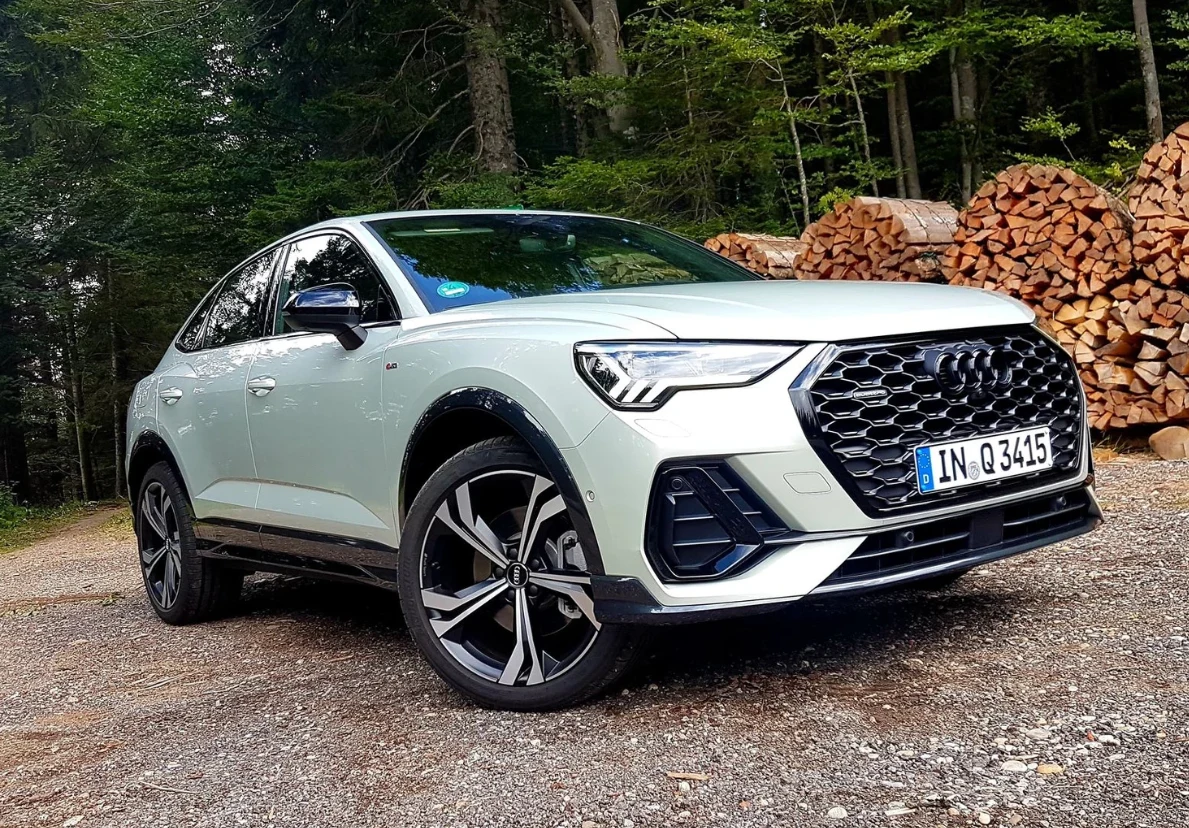 Nowoczesny samochód Audi SUV w srebrnym kolorze z wyraźnym, charakterystycznym grillem i sportowymi felgami stoi na parkingu leśnym, w tle widoczne stosy ułożonego drewna i otaczające drzewa.