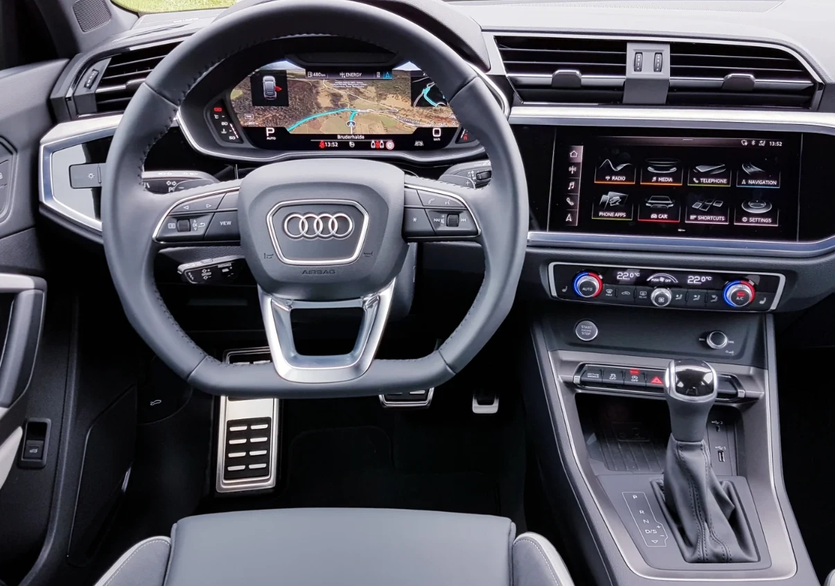 kokpit samochodu marki Audi z cyfrowym zestawem wskaźników, dużym ekranem systemu multimedialnego po prawej stronie, wielofunkcyjną kierownicą i automatyczną skrzynią biegów; widoczne nowoczesne wykończenie i detale wnętrza