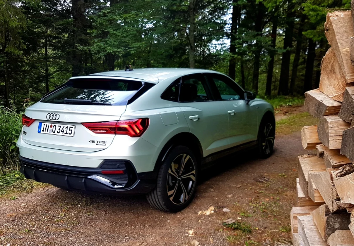 biała Audi Q3 Sportback zaparkowana na leśnej ścieżce, otoczona gęstymi drzewami, z prawej strony widoczny stos przeciętych, ułożonych kawałków drewna