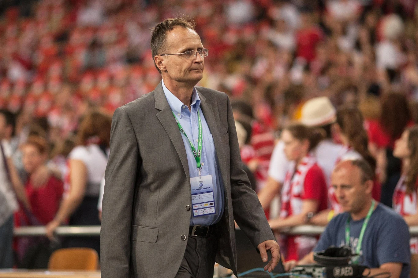 Mężczyzna w garniturze z identyfikatorem na szyi idzie przed tłumem ludzi na trybunach, w tle osoby ubrane w biało-czerwone stroje, sugerujące wydarzenie sportowe.