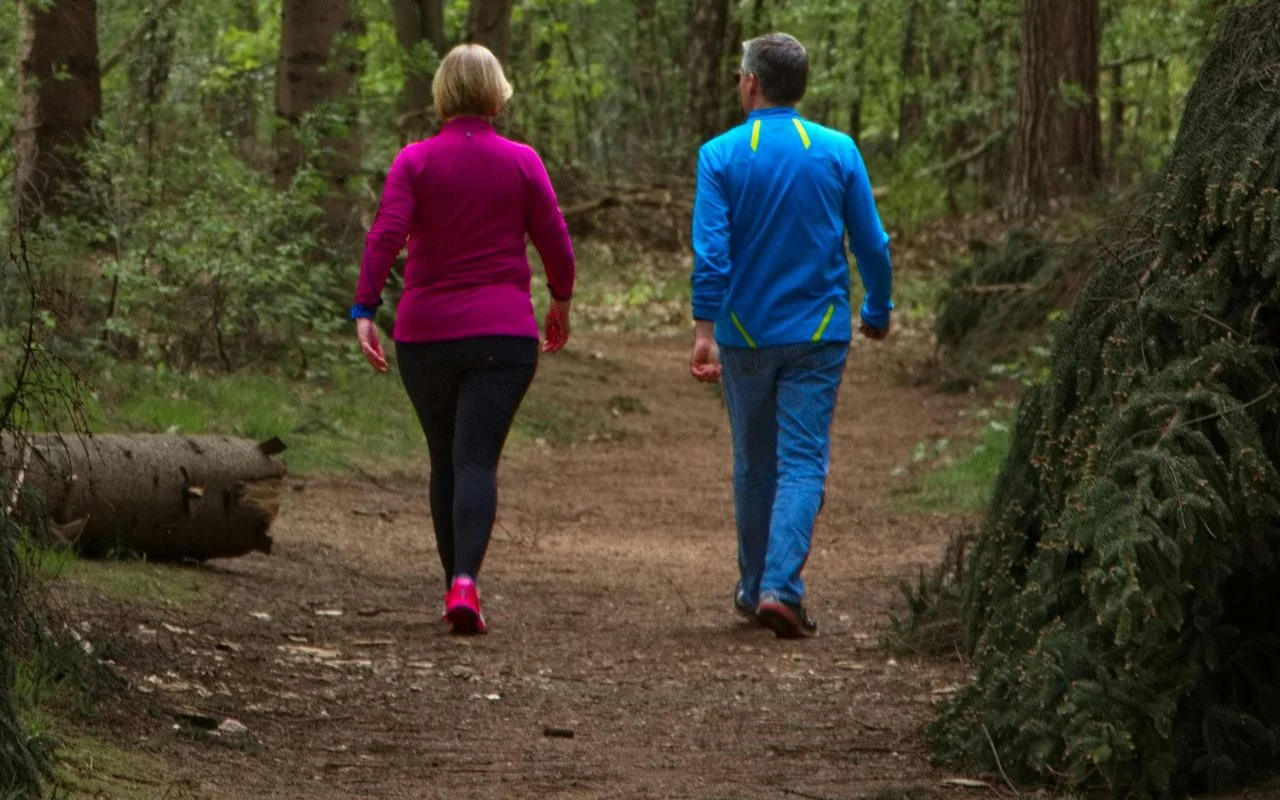 dwie osoby w sportowych ubraniach spacerują leśną ścieżką otoczoną drzewami i zielenią