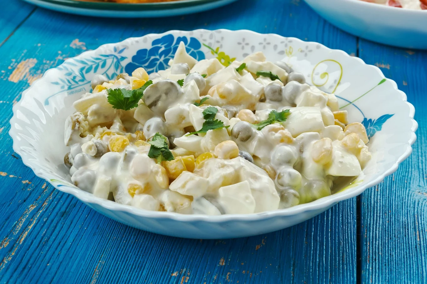 Prosta i sycąca sałatka, którą robi się w kilka chwil Sałatka z groszkiem, kukurydzą, ogórkiem kiszonym, jajkiem i majonezem w białej misce na niebieskim stole