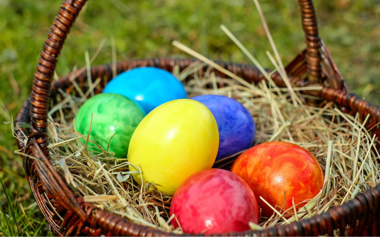 Koszyk z wikliny wypełniony sianem, w którym leży sześć kolorowych pisanek: żółta, niebieska, zielona, czerwona, pomarańczowa i różowa.