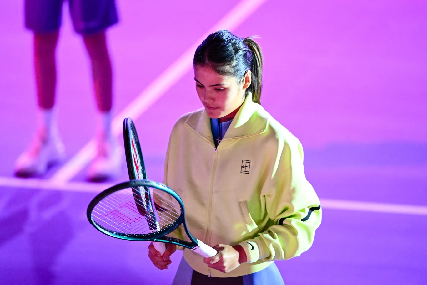 młoda kobieta w żółtej sportowej kurtce trzymająca dwa rakiety tenisowe na oświetlonym różowo-fioletowym korcie