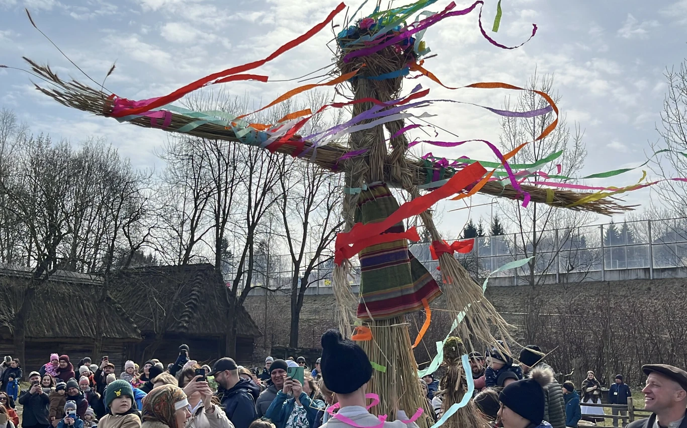 duża kukła wykonana ze słomy i ozdobiona kolorowymi wstążkami unosi się nad tłumem ludzi zgromadzonych podczas wiosennego obrzędu, w tle widać nagie drzewa oraz fragment drewnianych budynków