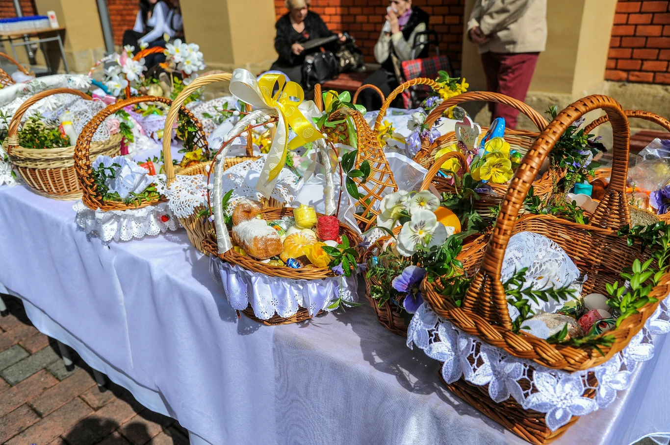 Kosze wiklinowe ozdobione białymi serwetkami, żółtymi i białymi wstążkami oraz świeżą zielenią, wypełnione tradycyjnymi pokarmami wielkanocnymi, ustawione na białym obrusie na zewnątrz, w tle osoby siedzące przy murze budynku.