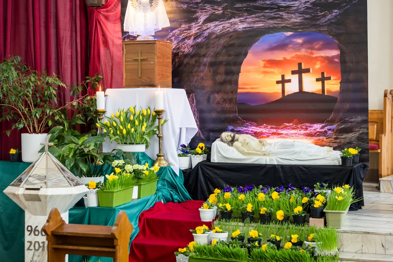 kompozycja Grobu Pańskiego w kościele z figurą leżącego Jezusa, otoczona licznymi żółtymi i fioletowymi kwiatami oraz zielonymi roślinami; w tle dekoracja przedstawiająca trzy krzyże na wzgórzu przy zachodzie słońca