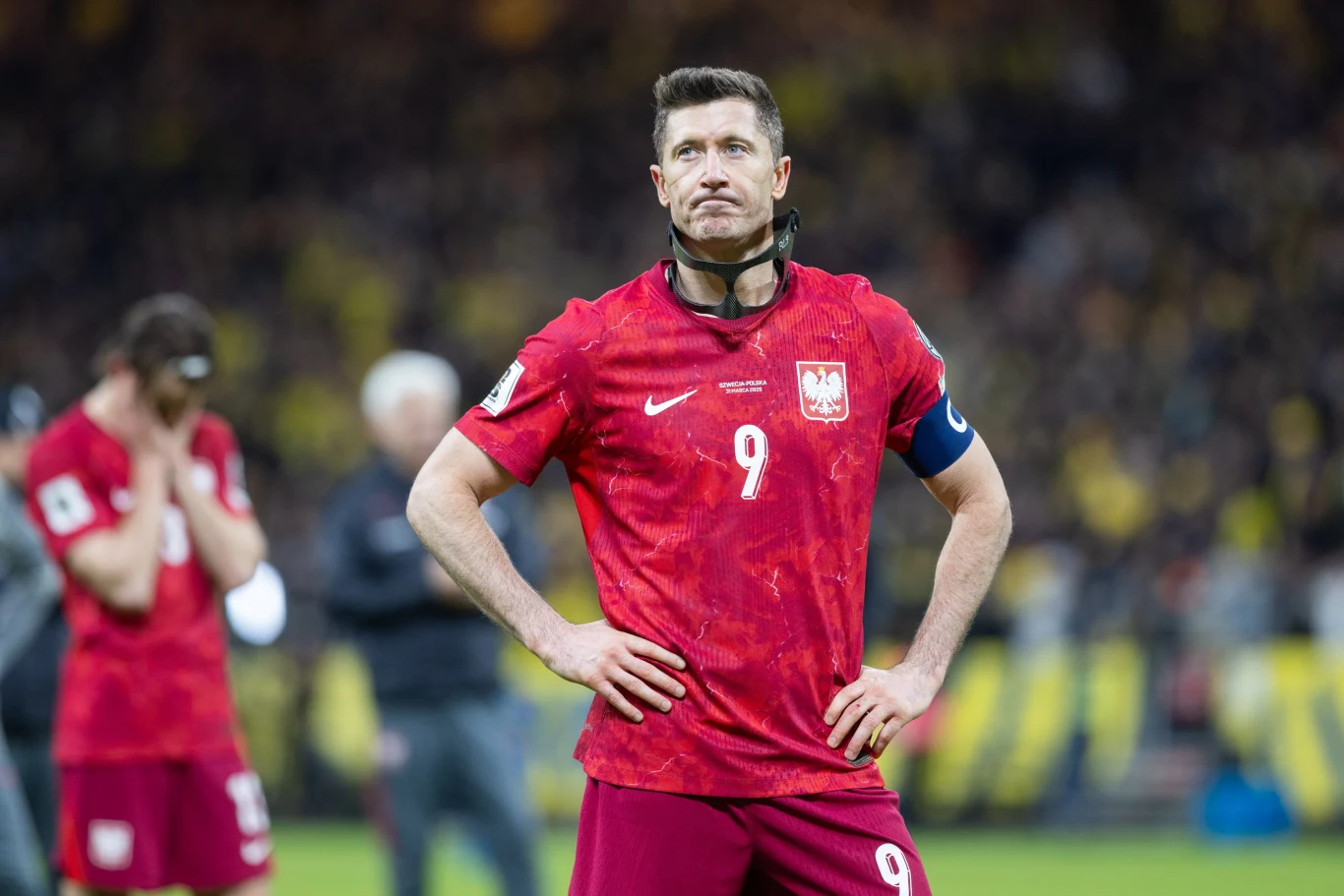 Robert Lewandowski po meczu ze Szwecją piłkarz w czerwonej koszulce z numerem 9 i opaską kapitańską stoi na boisku z rękami na biodrach, skupiony, w tle rozmyci zawodnicy i kibice
