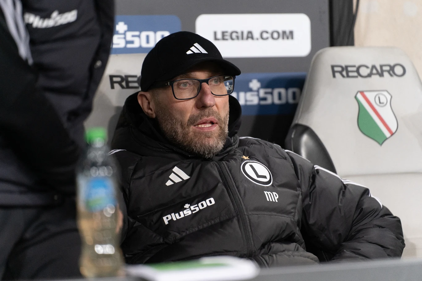 mężczyzna w okularach i czarnej czapce siedzący na ławce trenerskiej Legii Warszawa, ubrany w kurtkę z klubowym herbem, za nim widoczny znak Recaro oraz logo Legii