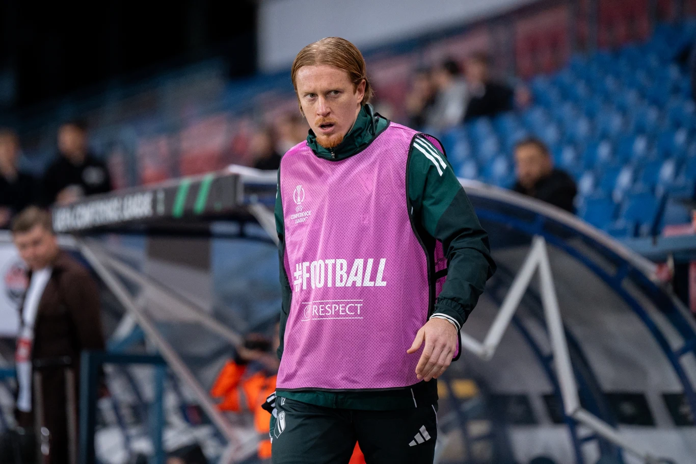 piłkarz w zielonym stroju z różową kamizelką treningową z napisem '#FOOTBALL RESPECT', stojący przy ławce rezerwowych na stadionie z pustymi niebieskimi krzesełkami w tle