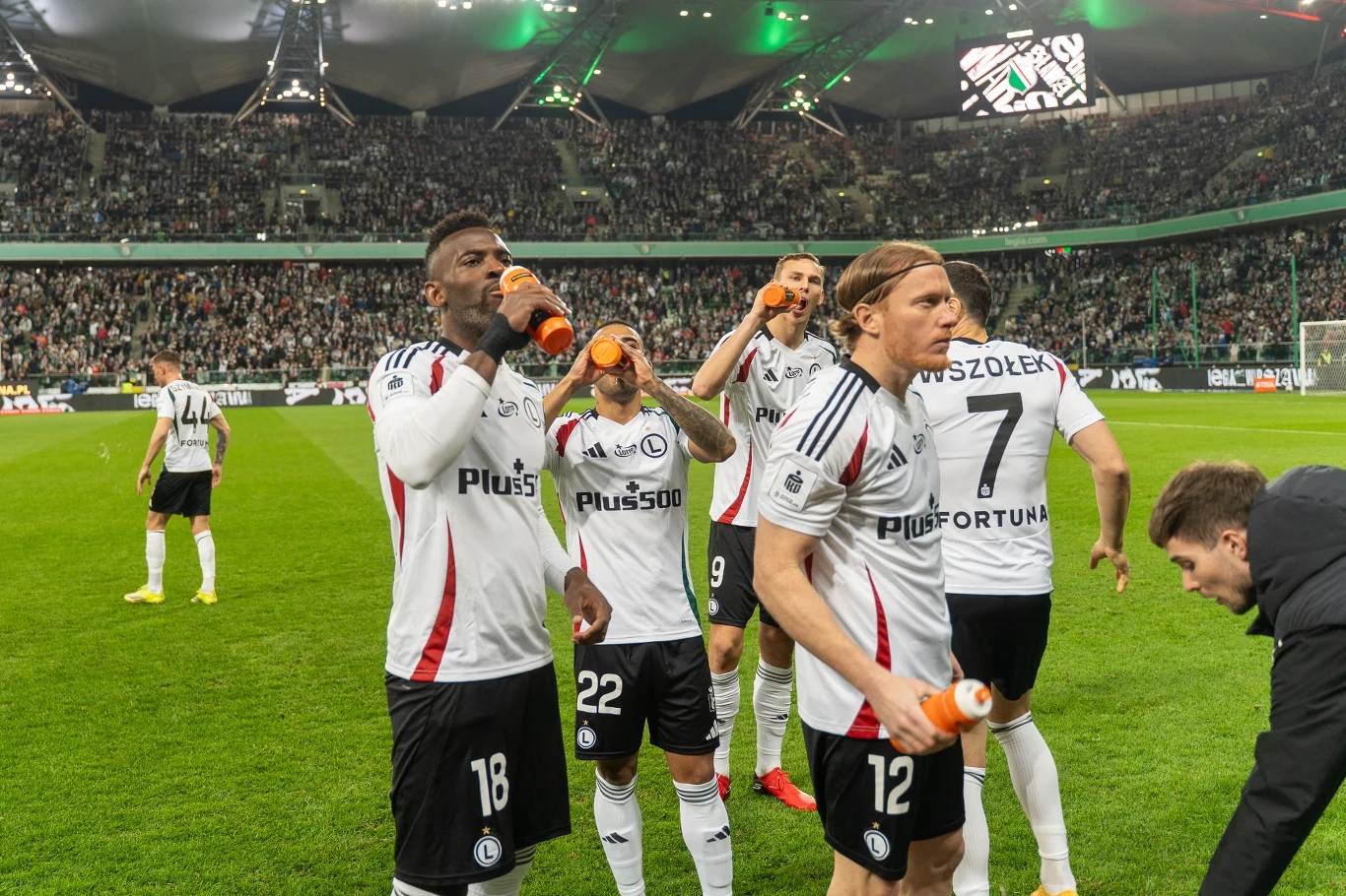 piłkarze drużyny w biało-czarnych strojach odpoczywają na murawie stadionu, pijąc napoje izotoniczne podczas przerwy meczu; trybuny wypełnione kibicami, intensywne światło stadionowe