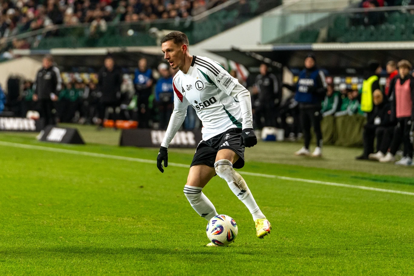 piłkarz w białym stroju z piłką przy nodze podczas meczu na stadionie, otoczony przez kibiców i innych zawodników w tle