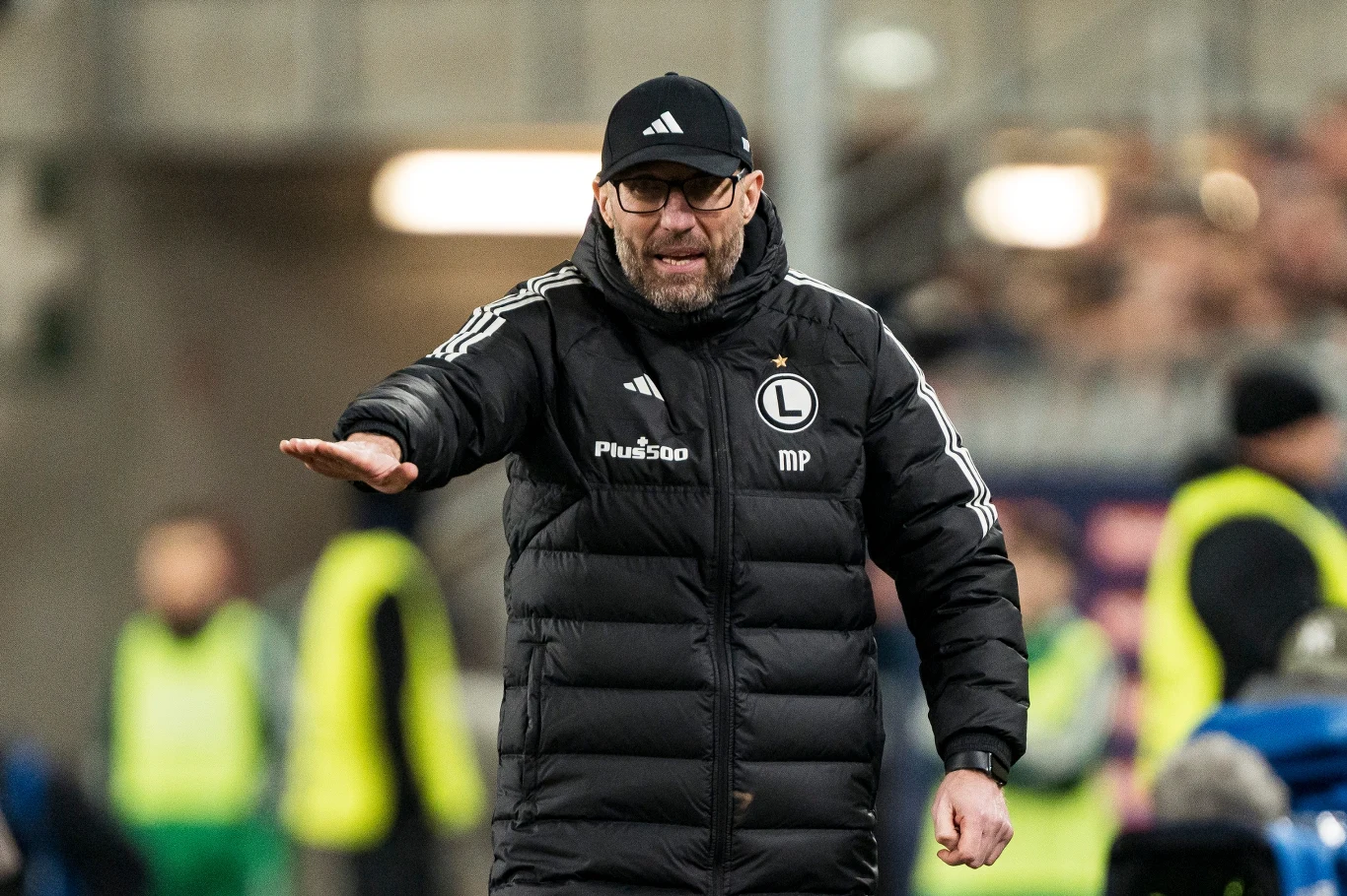 trener w czarnej kurtce z logotypem Legii Warszawa i czapce z daszkiem wydaje gestem ręki polecenie podczas meczu, w tle rozmyte sylwetki ludzi oraz stadionowe trybuny