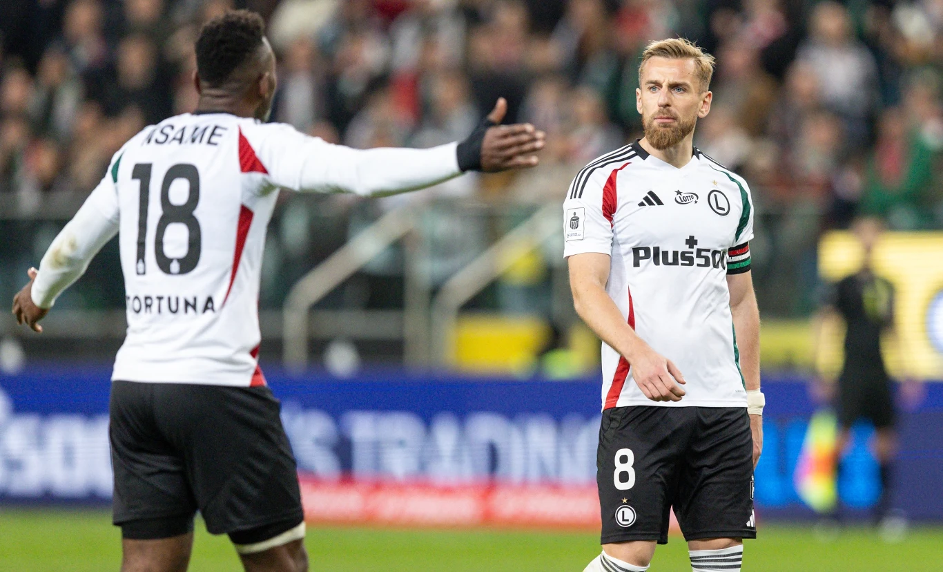 Dwóch piłkarzy drużyny Legia Warszawa w stroju meczowym podczas spotkania na boisku – jeden z nich z wyciągniętymi ramionami w kierunku drugiego, który patrzy przed siebie z poważnym wyrazem twarzy.