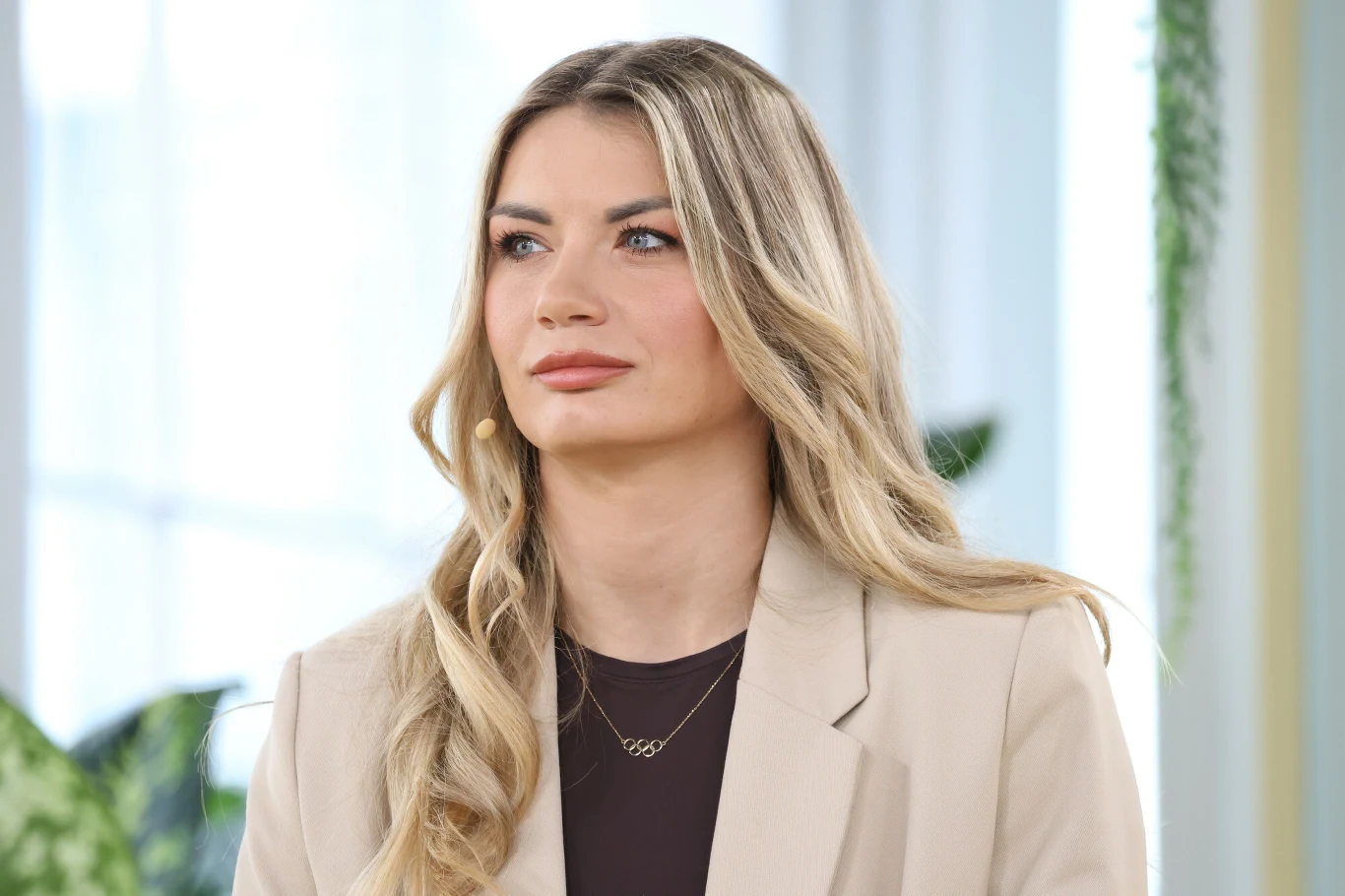 Kobieta o długich blond włosach, ubrana w jasny żakiet i ciemną bluzkę, z delikatnym makijażem oraz złotym naszyjnikiem, stoi w jasnym, nowoczesnym wnętrzu z zielonymi roślinami w tle.