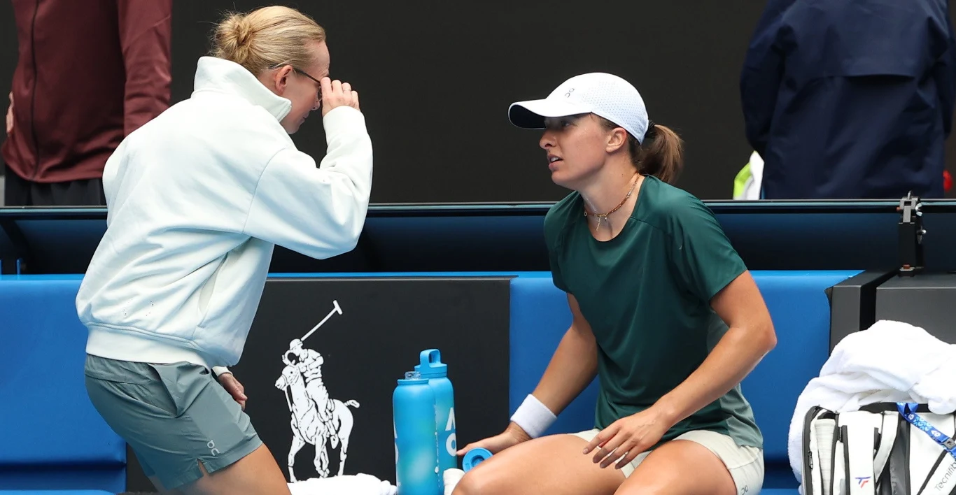 Tenisistka w zielonej koszulce i białej czapce siedzi na ławce z rakietą oraz butelką wody obok siebie, rozmawiając z osobą w jasnej bluzie, pochylającą się do przodu.