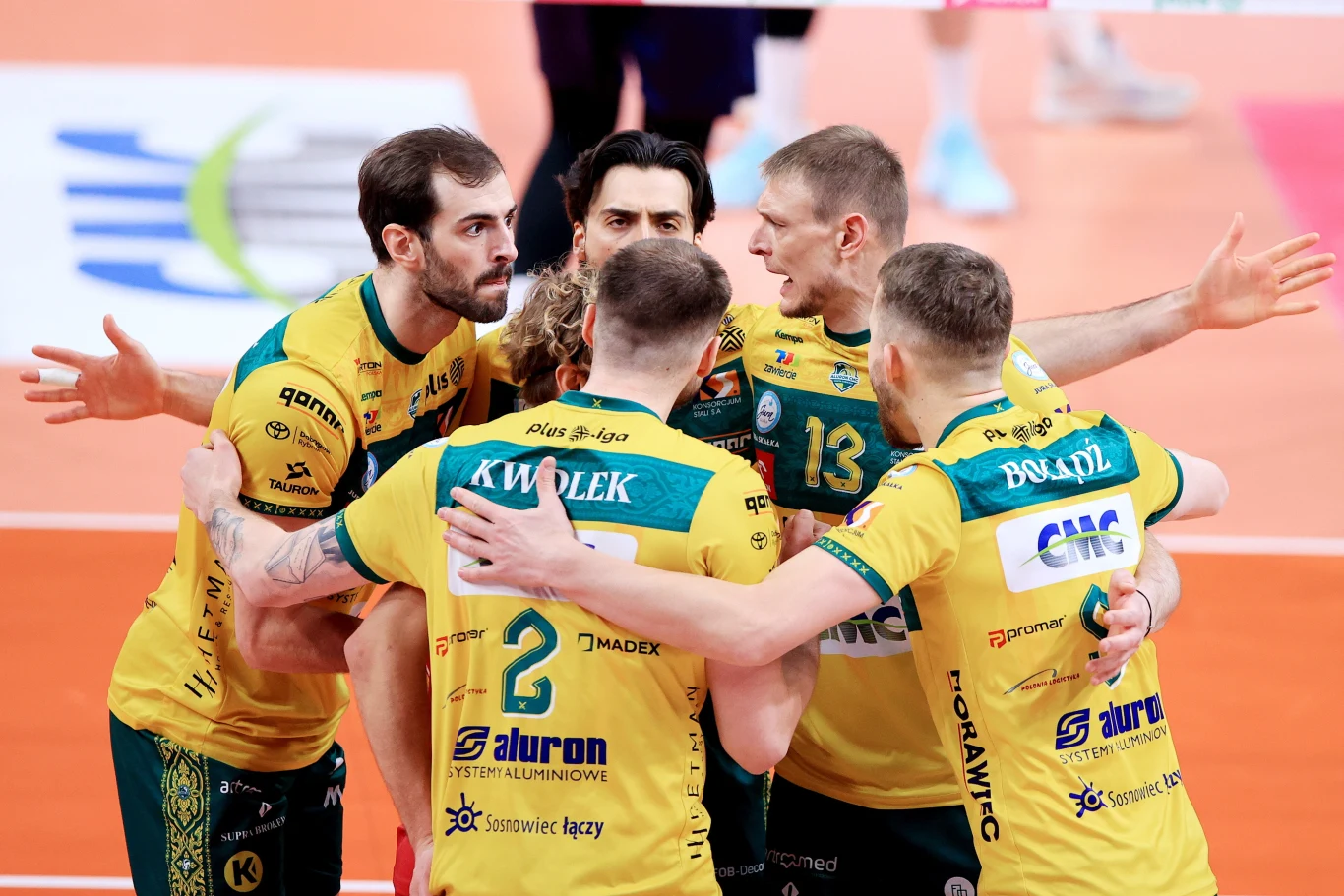 Grupa siatkarzy w żółto-zielonych strojach świętuje zdobycie punktu, wspólnie przytulając się na boisku do siatkówki, w tle widoczne białe linie wyznaczające pole gry.