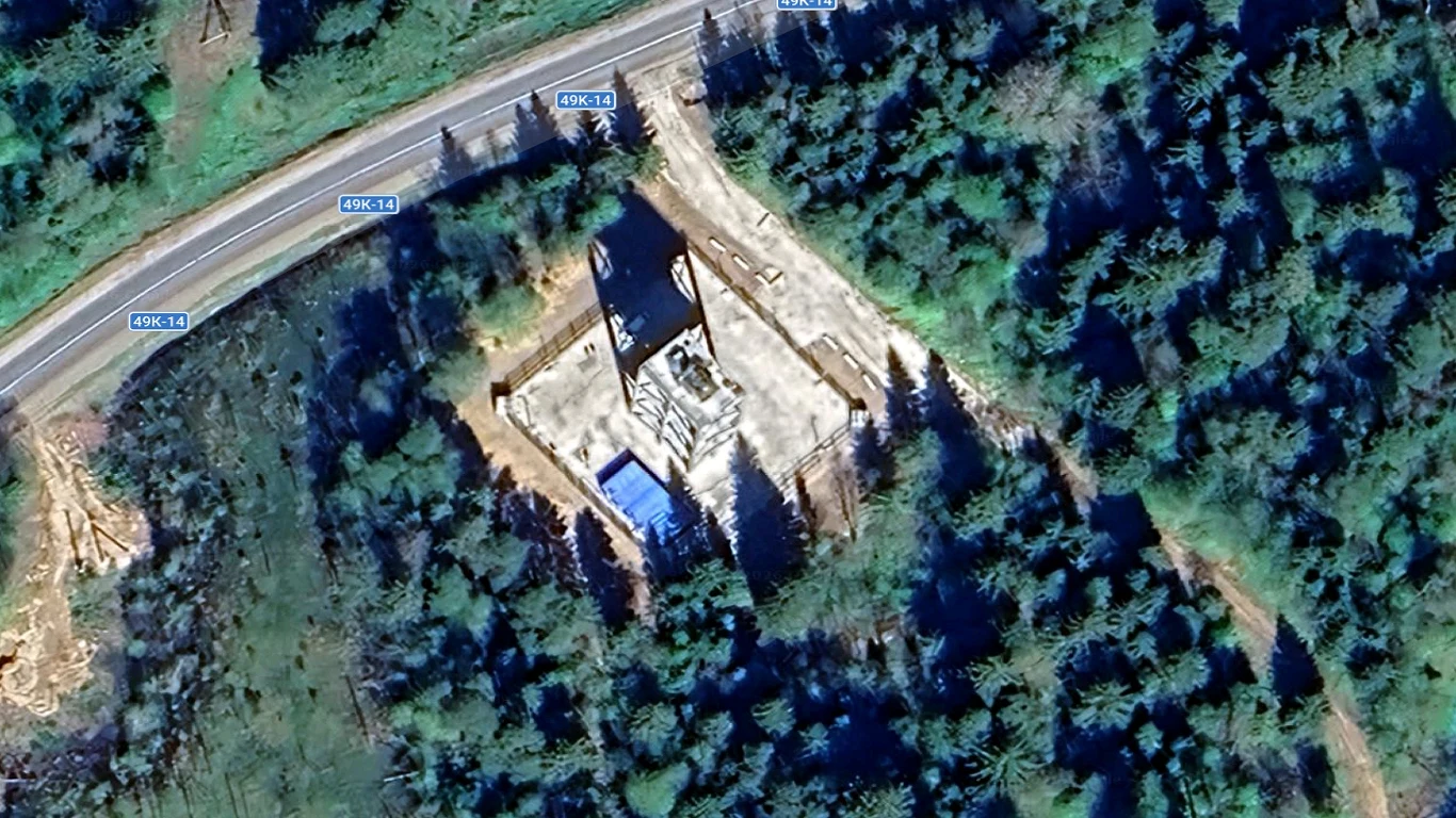 Budowa domu w stanie surowym otwartym na prostokątnej działce w otoczeniu gęstego lasu, obok biegnie asfaltowa droga oznaczona tabliczkami z numerem DK16.