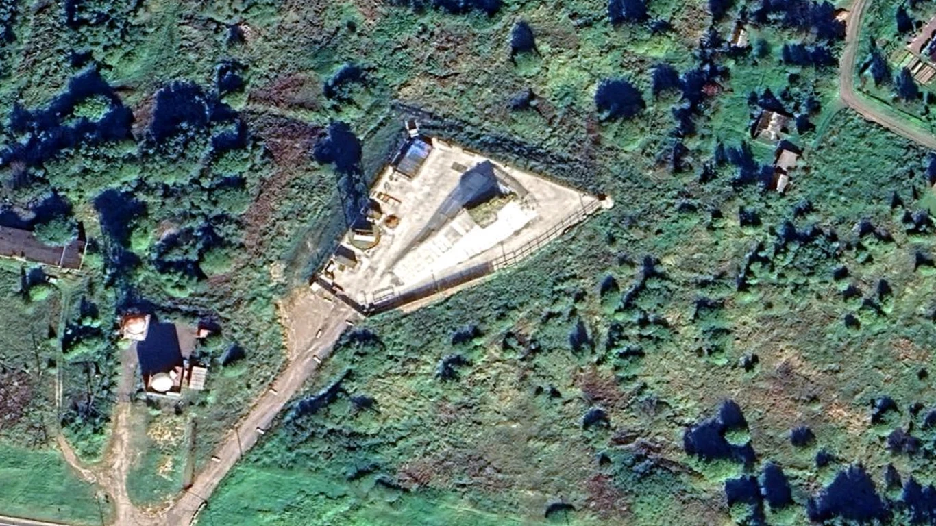 trójkątny budynek o jasnym dachu z centralnie umieszczoną ciemniejszą konstrukcją, otoczony zielenią i kilkoma niewielkimi zabudowaniami po bokach