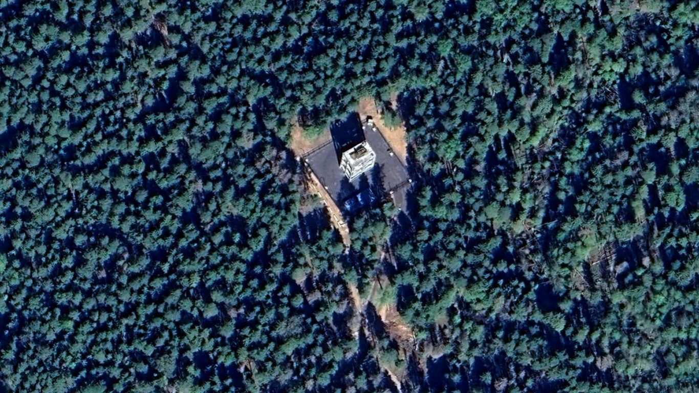 prostokątny budynek otoczony gęstym, zielonym lasem iglastym, z widocznymi drogami lub ścieżkami prowadzącymi do obiektu