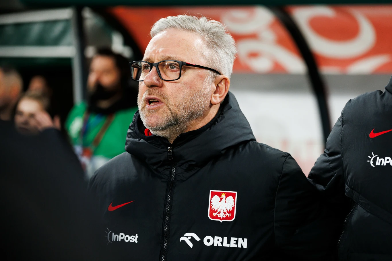 Mężczyzna w okularach, ubrany w kurtkę z godłem Polski i logotypami sponsorów, stoi na tle stadionu sportowego i wygląda na skupionego.