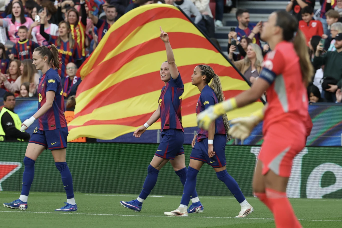 piłkarki drużyny FC Barcelona w granatowo-bordowych strojach świętują na murawie stadionu, jedna z nich z uniesioną ręką wskazuje w górę, w tle tłum kibiców oraz duża katalońska flaga w żółto-czerwone pasy