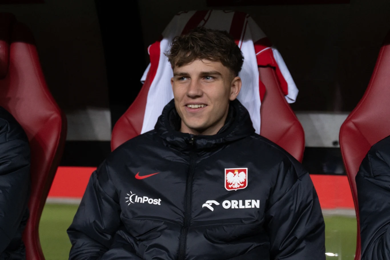 młody mężczyzna w kurtce z emblematem reprezentacji Polski i logotypami sponsorów siedzi na czerwonym stadionowym fotelu, w tle rozwieszona biało-czerwona koszulka