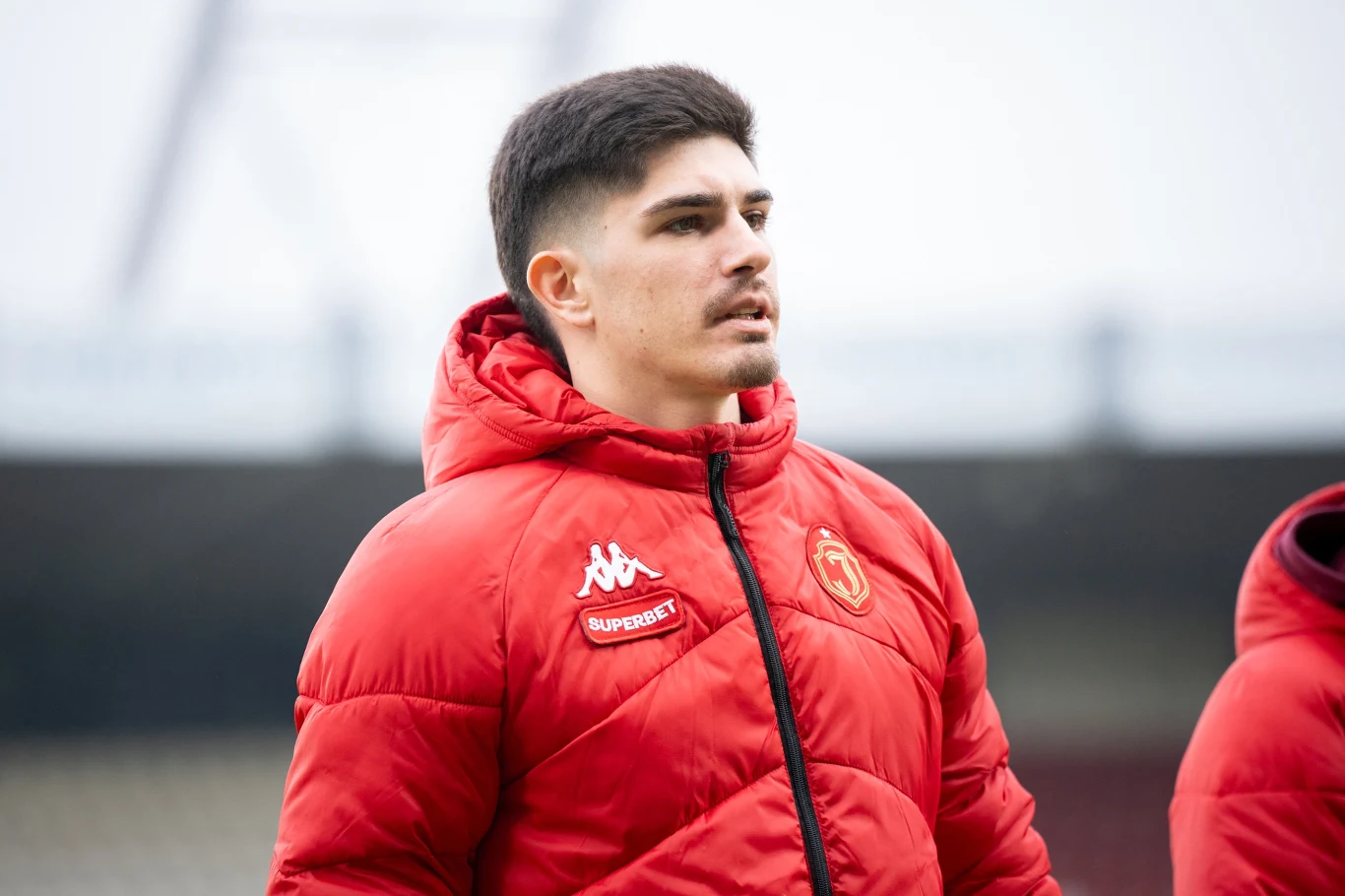 młody mężczyzna w czerwonej kurtce sportowej z logo Kappa i emblematem klubu, stojący na tle rozmytego stadionu