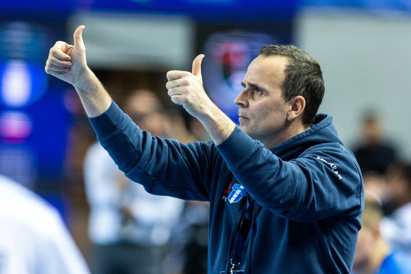 mężczyzna ubrany w niebieską sportową bluzę pokazuje oba kciuki w górę w geście aprobaty, znajdując się w sportowej hali