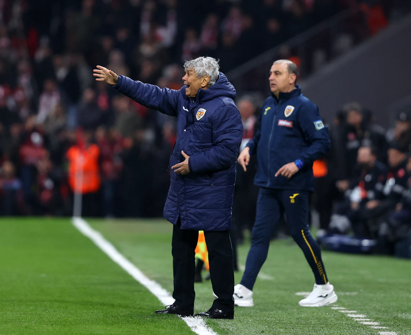 trener w granatowej kurtce energicznie gestykuluje przy linii bocznej boiska podczas meczu piłkarskiego, obok niego stoi drugi członek sztabu w dresie