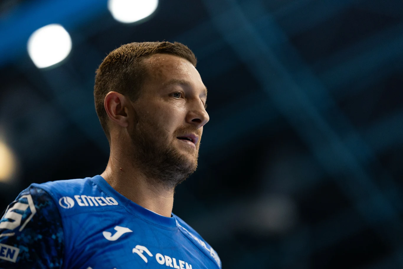 mężczyzna w sportowej koszulce z logotypami sponsorów na tle rozmytego wnętrza hali sportowej, skupiony wyraz twarzy podkreśla atmosferę zawodów