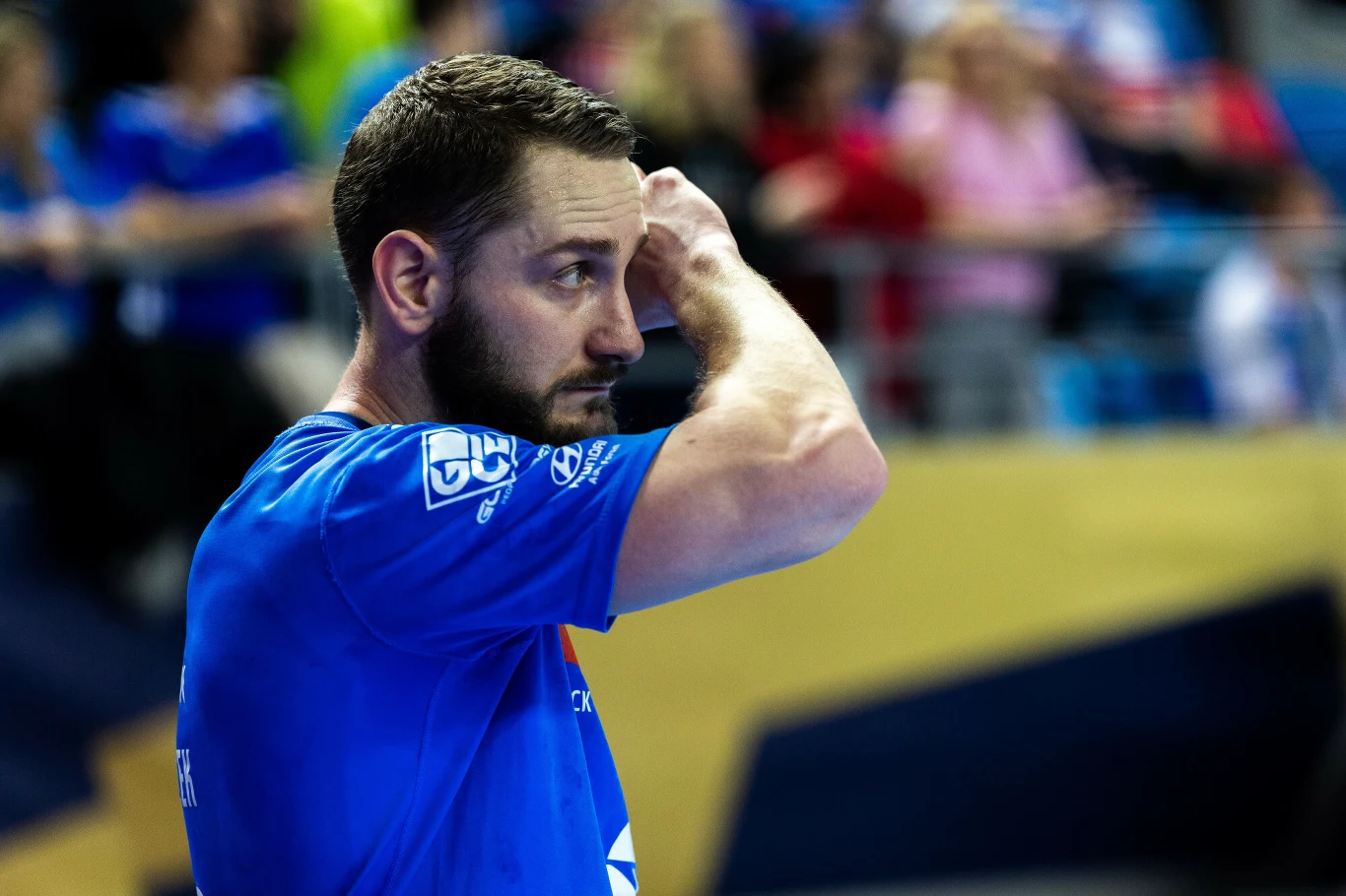 sportowiec w niebieskiej koszulce poprawia włosy, za nim rozmazana publiczność na trybunach