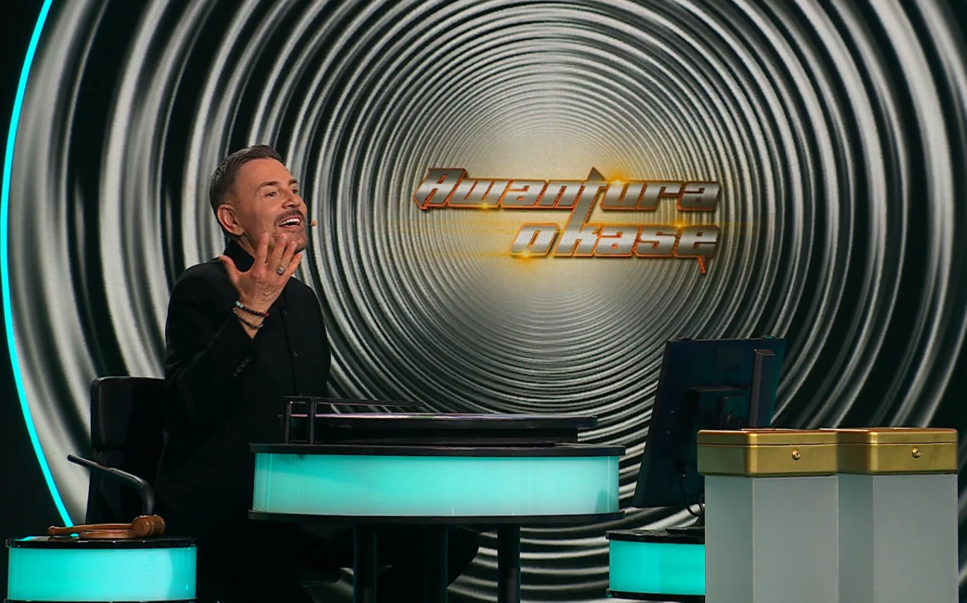 Krzysztof Ibisz za nowoczesnym biurkiem, gestykulujący, z logo programu "Awantura o kasę".