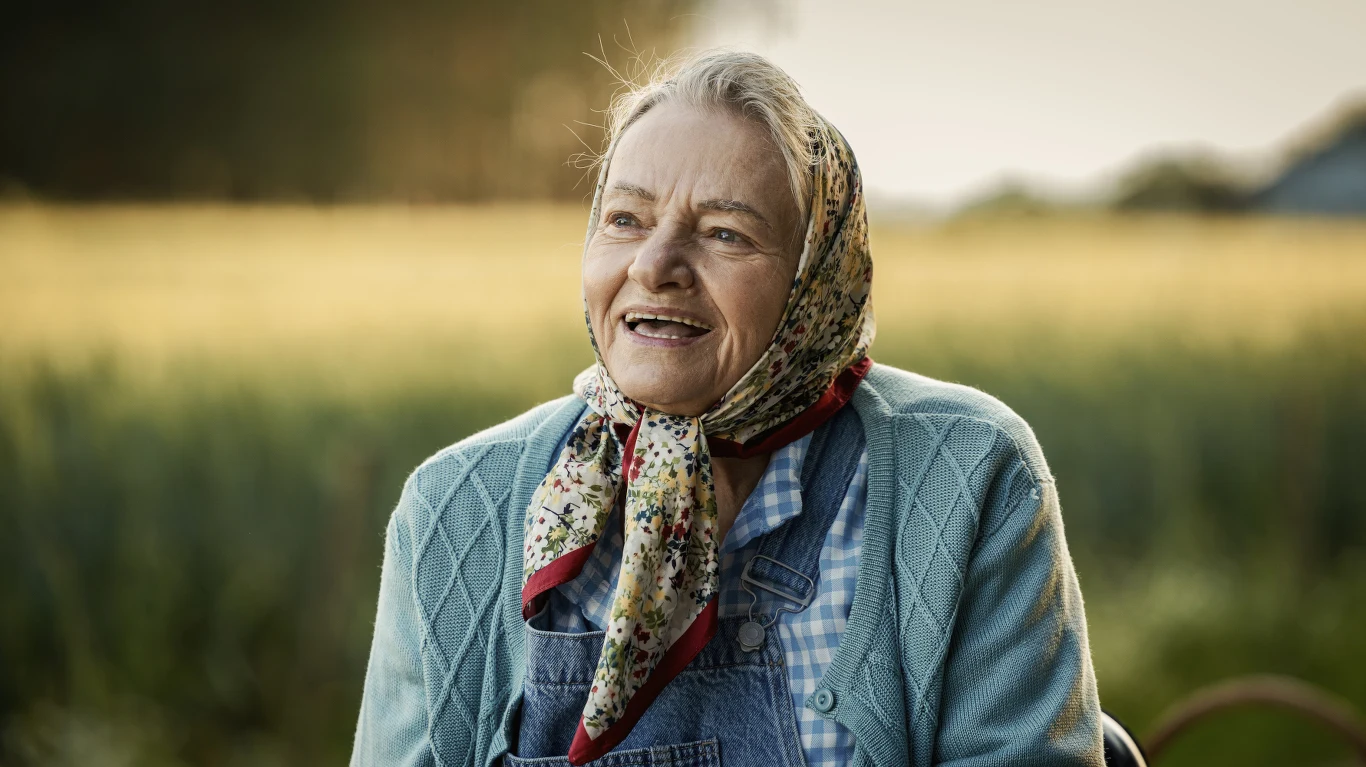 Starsza kobieta z uśmiechem na twarzy, ubrana w sweter, koszulę w kratę i kolorową chustkę na głowie, siedzi na tle rozmytego, słonecznego pola.