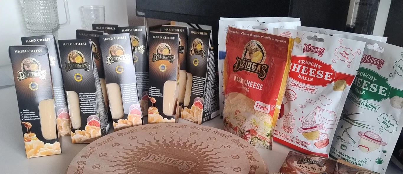 Różnorodne opakowania serów przekąskowych i twardych w pudełkach i saszetkach ustawione obok siebie na blacie, na pierwszym planie okrągłe pudełko z ozdobnym wzorem, dominują produkty marki Džiugas oraz chrupiące przekąski serowe w różnych smakach.