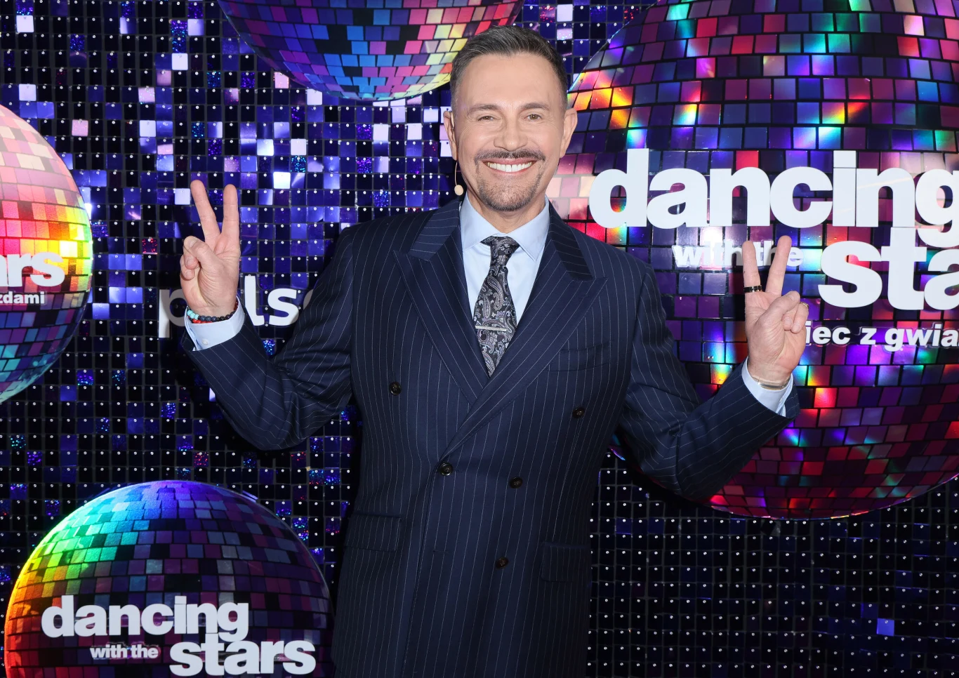 Mężczyzna w eleganckim garniturze w paski stoi na tle kolorowych, lustrzanych kul oraz logo programu 'Dancing with the Stars', uśmiecha się i pokazuje dłońmi znak pokoju po obu stronach.