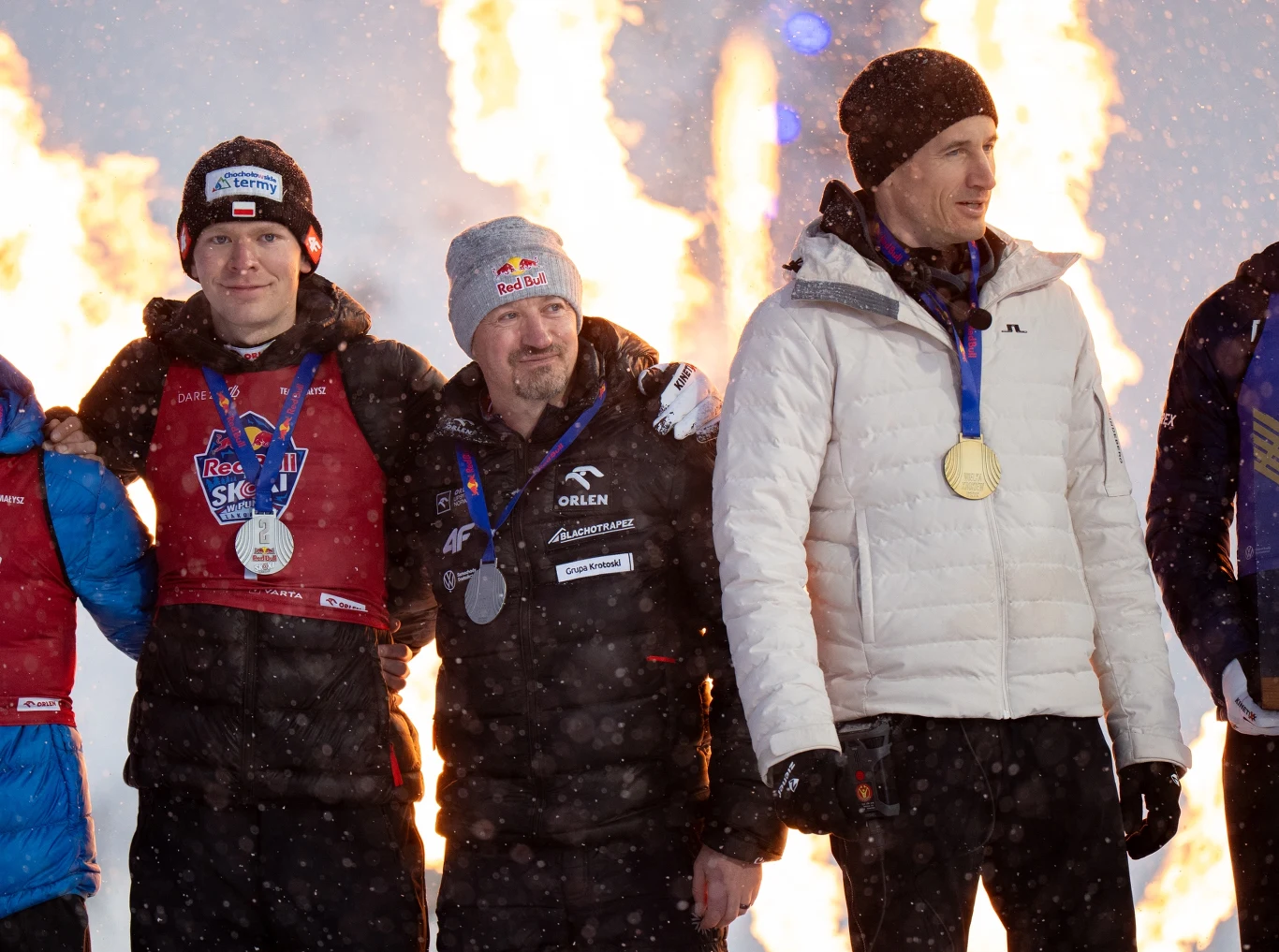 Trzech mężczyzn ze srebrnymi i złotymi medalami stoi ramię w ramię na podium w zimowej scenerii, w tle płomienie fajerwerków i padający śnieg, atmosfera zwycięstwa i sportowej rywalizacji.