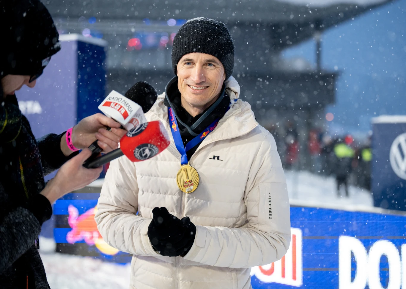 Mężczyzna w białej kurtce i czarnej czapce stoi na śniegu, na szyi ma złoty medal i niebieską wstążkę, udziela wywiadu osobie trzymającej mikrofon z logo stacji telewizyjnej, w tle widoczne elementy scenografii i rozmyte postaci.