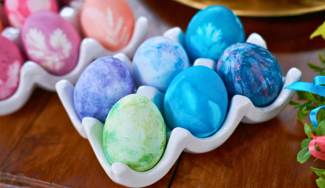 Techniki zdobienia pisanek były niezwykle różnorodne Kolorowe pisanki w pastelowych odcieniach zieleni, fioletu i niebieskiego ułożone w białym ceramicznym pojemniku na drewnianym stole, z widoczną częścią różowych pisanek w tle.