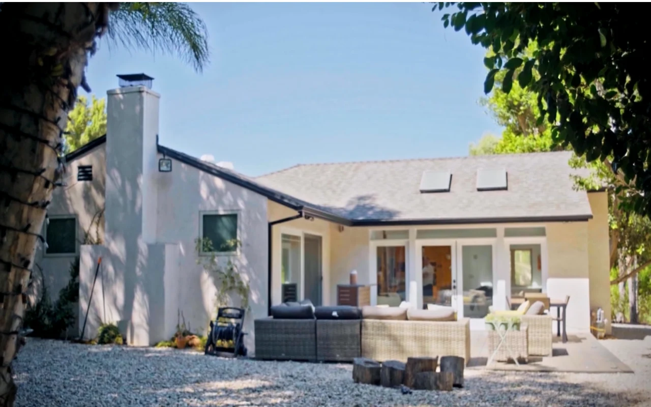 Nowoczesny, jasny dom jednorodzinny z dużymi oknami i tarasem, na którym ustawiono zestaw wypoczynkowy z szarych mebli ogrodowych oraz stolik z drewnianych pni, otoczony zielenią i cienistymi drzewami.