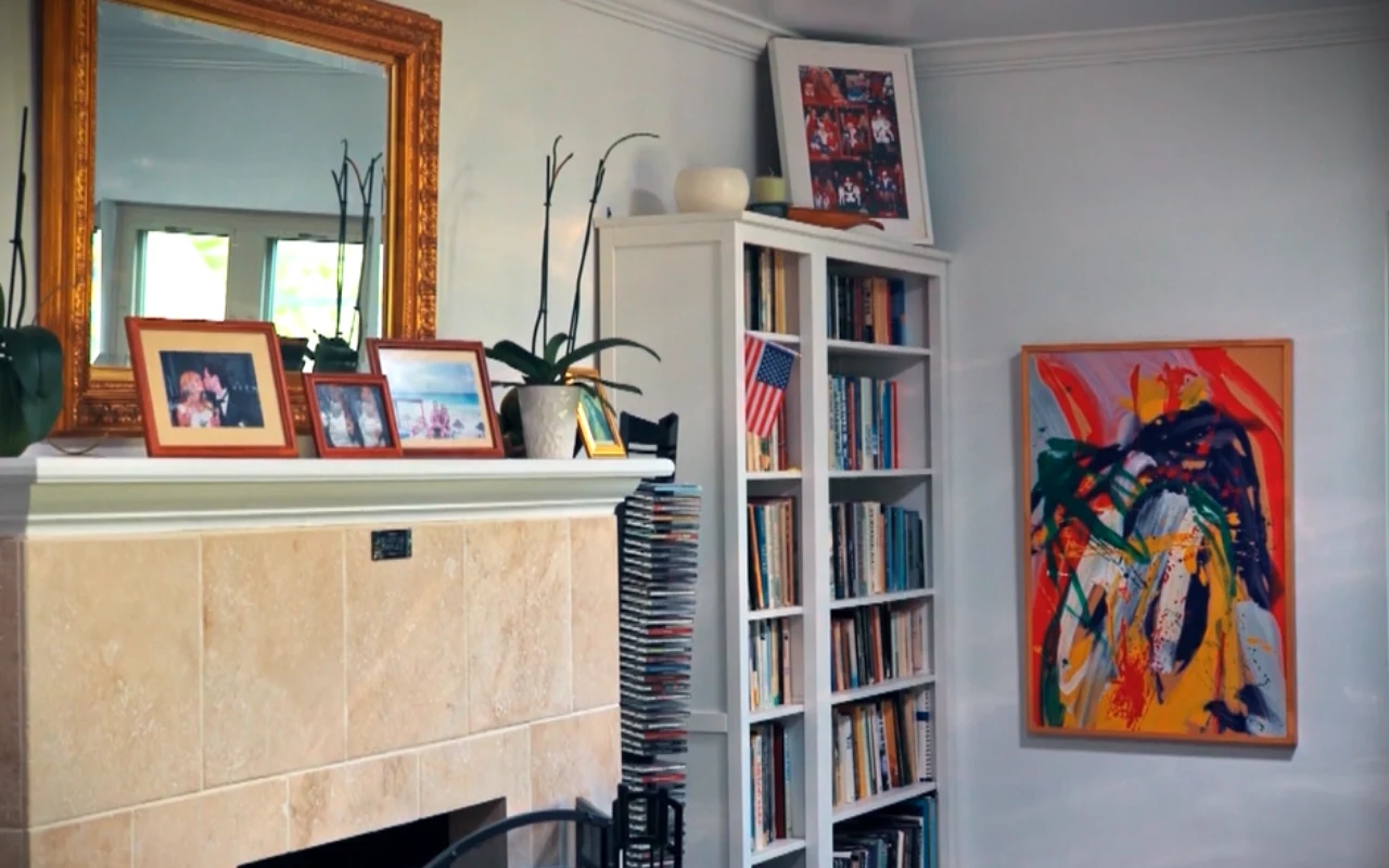 Kominek z jasnym marmurowym obramowaniem, na którym ustawione są rodzinne zdjęcia w ramkach i ozdobne rośliny, obok biała biblioteczka wypełniona książkami oraz kolorowy abstrakcyjny obraz na sąsiedniej ścianie.