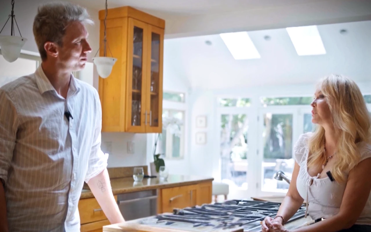 Dwoje dorosłych rozmawia przy kuchennej wyspie w nowoczesnej, jasnej kuchni z drewnianymi szafkami i dużymi oknami wpuszczającymi naturalne światło.