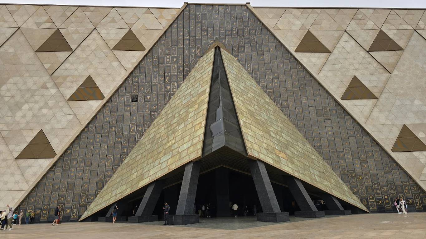 nowoczesny budynek z monumentalnym trójkątnym wejściem przypominającym piramidę, otoczony geometrycznymi zdobieniami ścian, z niewielką liczbą osób u podstawy konstrukcji