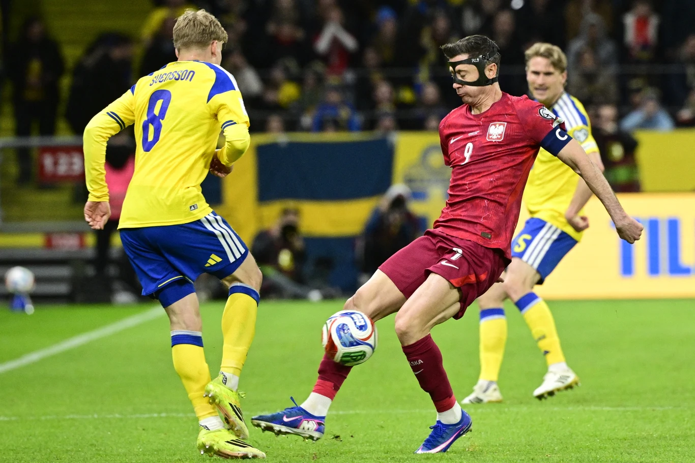 Robert Lewandowski w czerwonym stroju reprezentacji Polski prowadzi piłkę na boisku podczas meczu przeciwko Szwecji, naprzeciwko niego ustawia się zawodnik w żółto-niebieskim stroju z numerem 8.