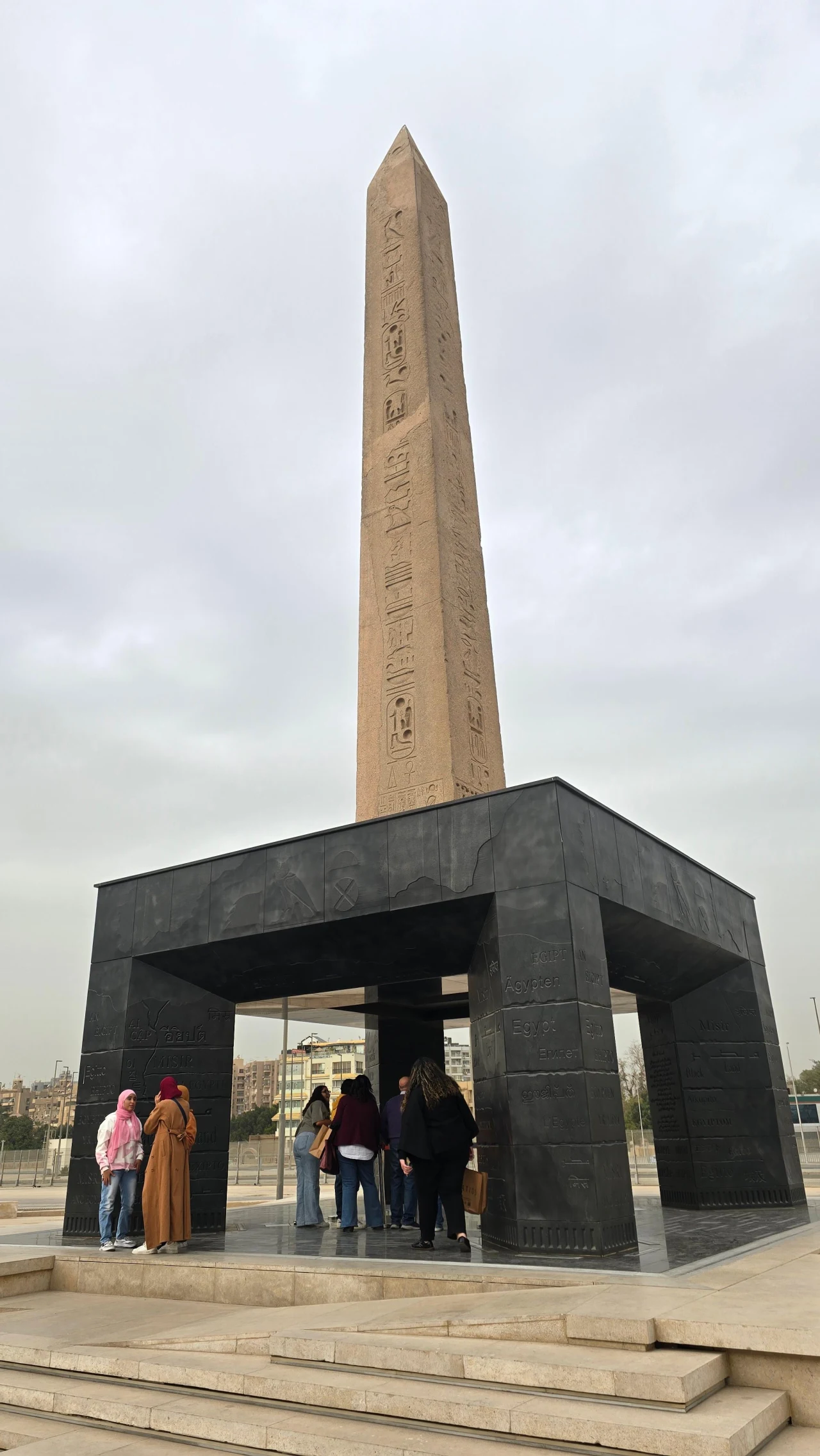 Wysoki obelisk z wyrytymi hieroglifami osadzony na czarnej, nowoczesnej konstrukcji, pod którą kilka osób przygląda się monumentowi.