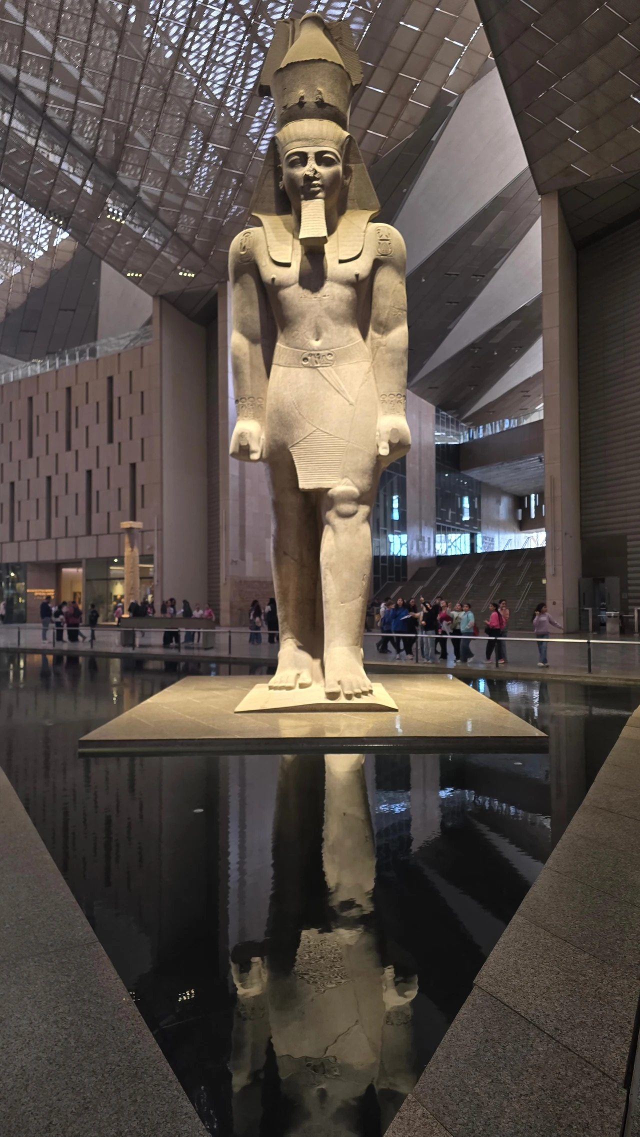 Monumentalny posąg faraona Ramzesa II ustawiony na środku atrium Wielkiego Muzeum Egipskiego, otoczony przez basen wodny odbijający jego sylwetkę, z grupą zwiedzających w tle.